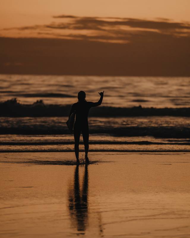 Surfer walking out of the sea at sunset, board under the arm, waving a shaka