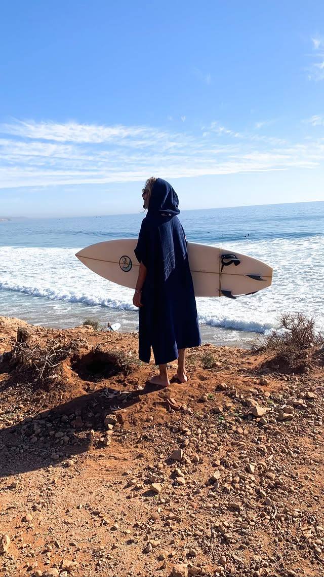 Surfer in a poncho holding a board on the cliff, looking at the waves