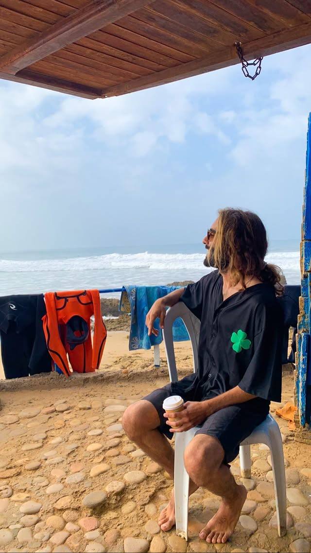 Surfer sitting with a coffee under a beach shelter, wetsuits drying, the waves behind