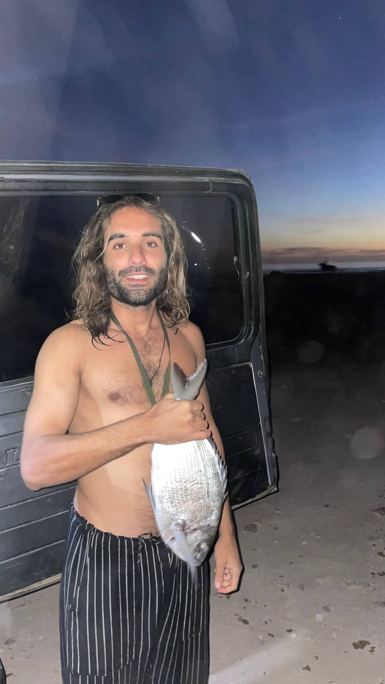 Smiling surfer holding a fish by the 4x4 at dusk