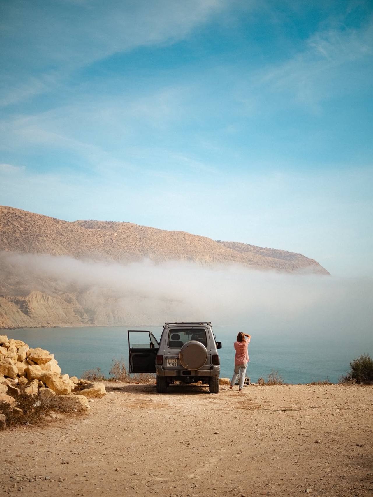A 4x4 parked on a clifftop above the sea, someone looking out at the bay, morning fog along the hills