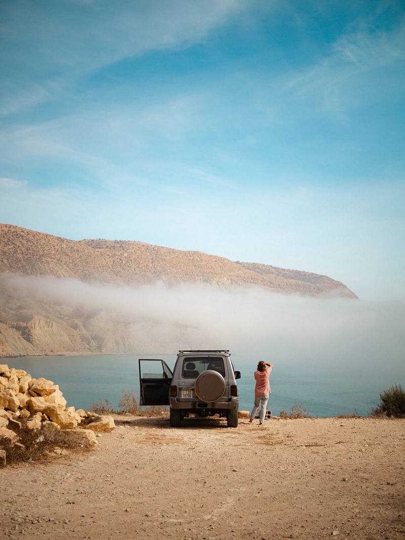 A 4x4 parked on a clifftop above the sea, someone looking out at the bay, morning fog along the hills